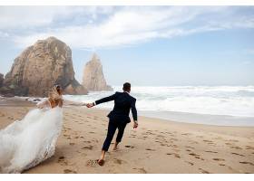 幸福的新婚夫妇牵着手在大西洋的海滩上奔跑_831683701