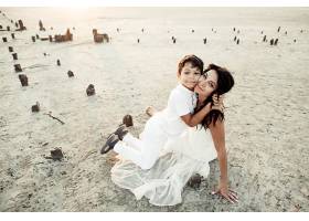 母子俩穿着白衣坐在沙滩上微笑着拥抱着_7249526