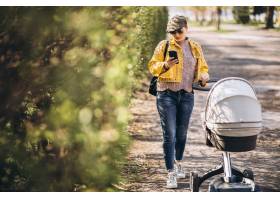 年轻的母亲推着婴儿车在公园里散步使用手_7870004